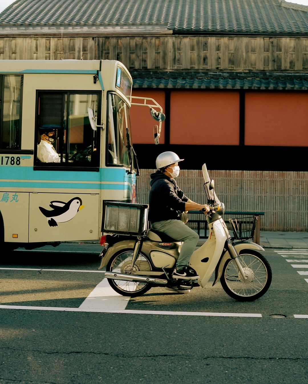 A man in a helmet and mask rides a cream-colored vintage moped on a city street, with a bus and traditional building in the background.