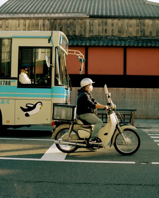 A man in a helmet and mask rides a cream-colored vintage moped on a city street, with a bus and traditional building in the background.