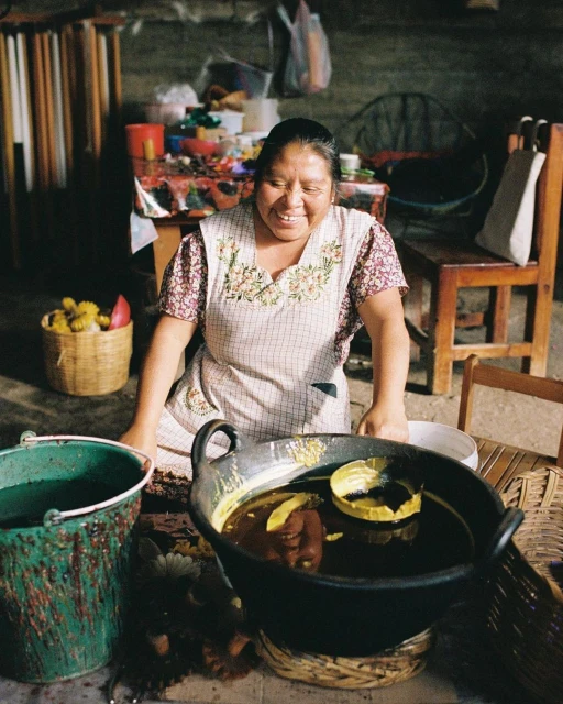 Smiling woman in apron prepares food in a large pot indoors, with flowers, a bucket, and kitchenware visible in a rustic setting.