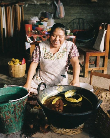 Smiling woman in apron prepares food in a large pot indoors, with flowers, a bucket, and kitchenware visible in a rustic setting.