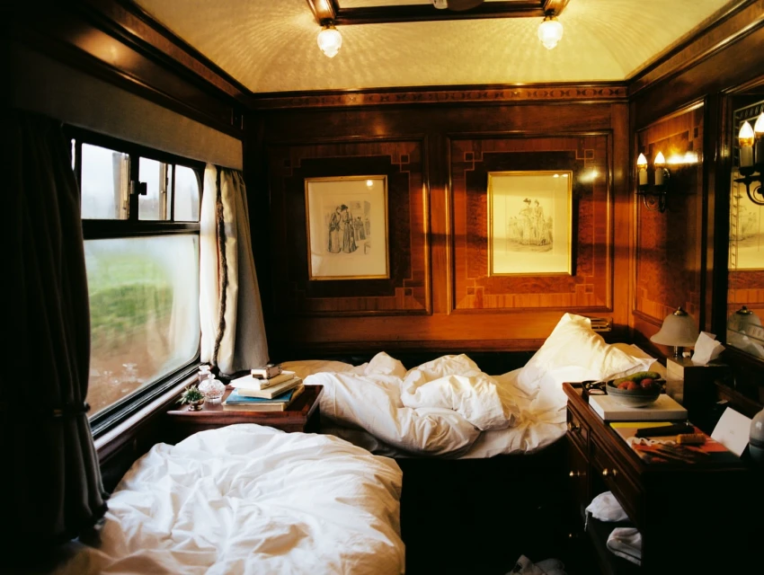 Inside a vintage train car, a double bed sits next to a window with curtains. Drawings adorn the wood-paneled walls, lit by sconces.