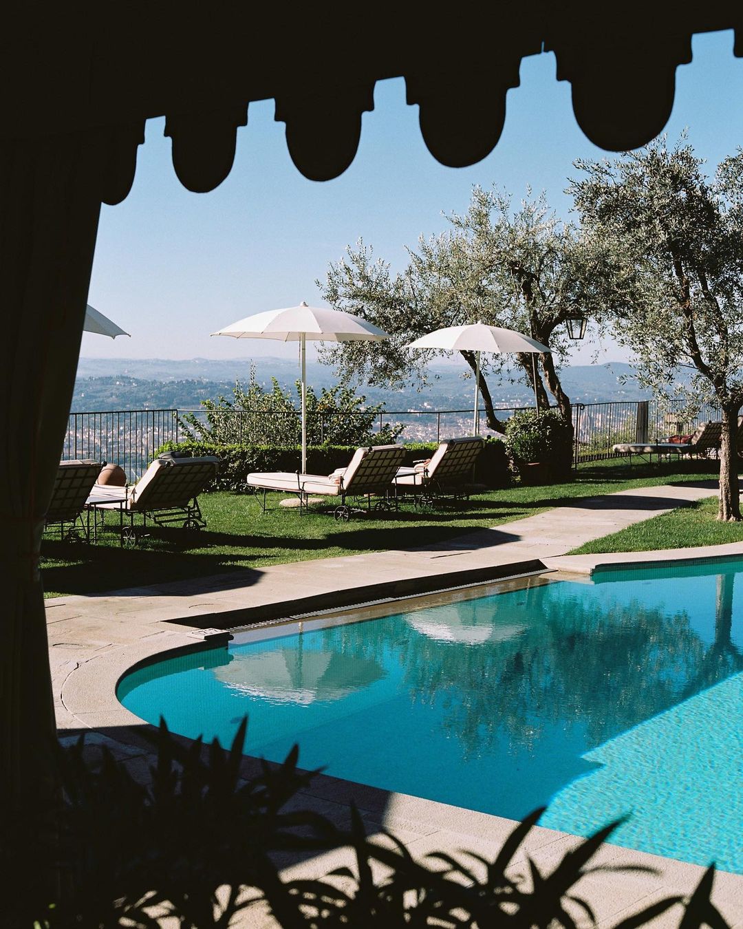 Outdoor swimming pool area with lounge chairs and umbrellas, framed by foliage and a building structure, with a view of distant hills.