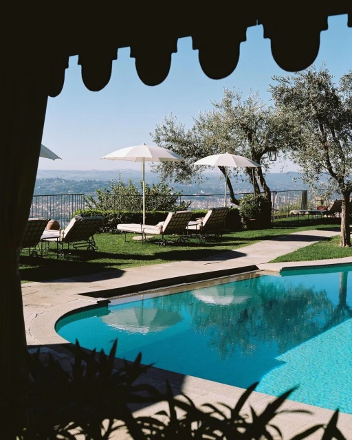 Outdoor swimming pool area with lounge chairs and umbrellas, framed by foliage and a building structure, with a view of distant hills.