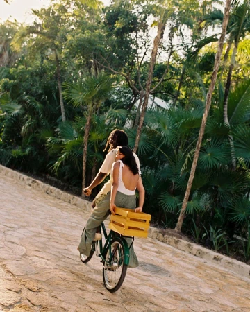 A couple riding a bicycle on a paved path lined with lush green vegetation; the woman carries a wooden crate on the back.
