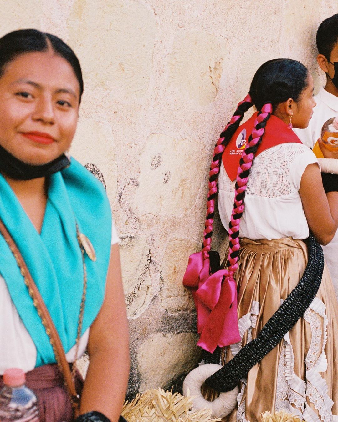 Two women in traditional dress stand near a textured wall, one with long braids and a colorful scarf.  Portrait orientation.