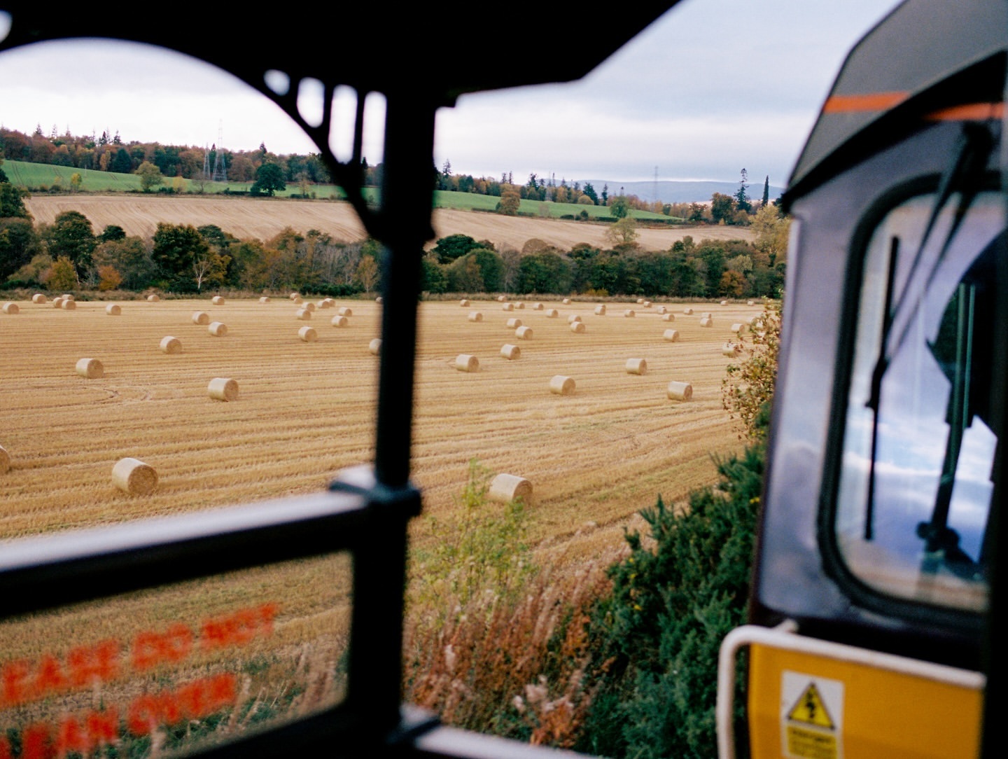 View of golden hay bales in a rural field from the window of a vehicle, possibly a train, with a partially visible black frame.