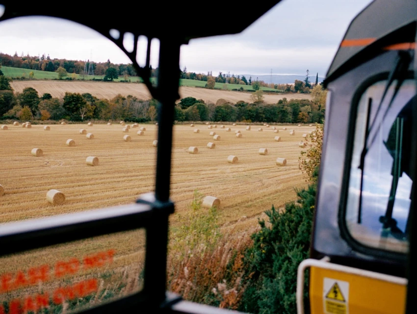 View of golden hay bales in a rural field from the window of a vehicle, possibly a train, with a partially visible black frame.