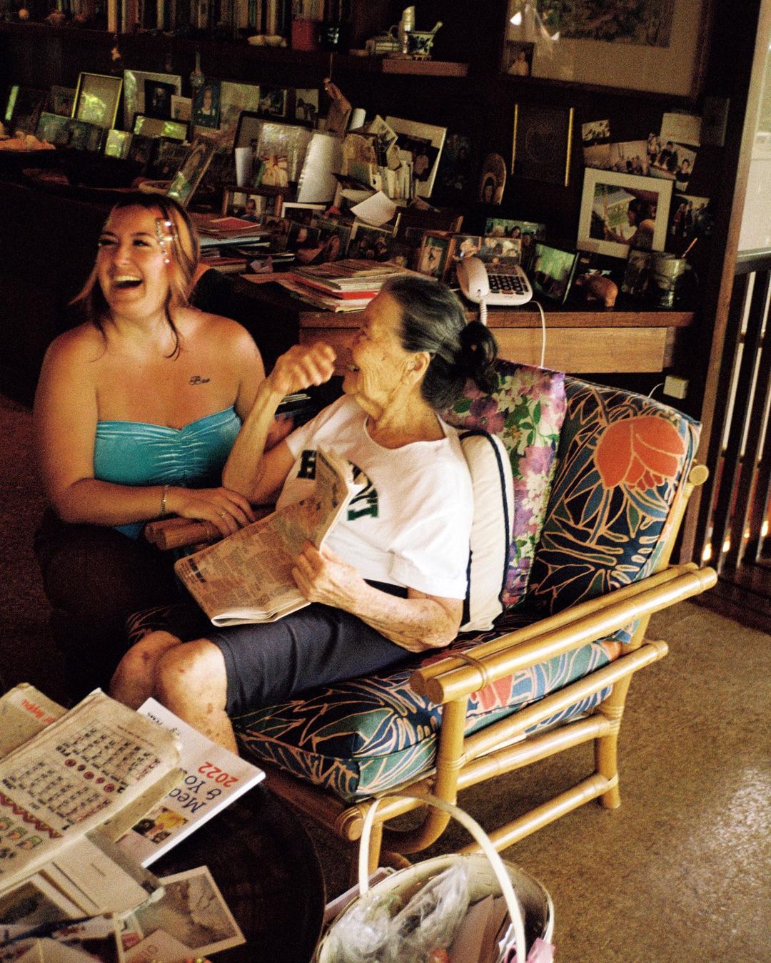 Two women share a lighthearted moment indoors, one in a turquoise top, the other seated in a floral chair, reading a newspaper, laughing.