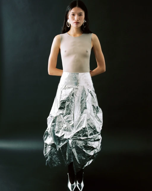Asian woman in silver foil skirt and neutral top against a black background, accessorized with hoop earrings and silver pointed heels.