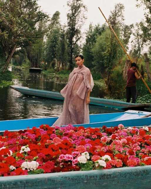 A woman in a sheer dress stands in a blue boat filled with red and pink flowers, another boat with a man visible behind.