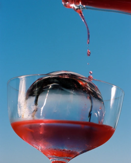 Red liquid being poured from a bottle into a clear wine glass with a sphere of ice, set against a bright blue background.