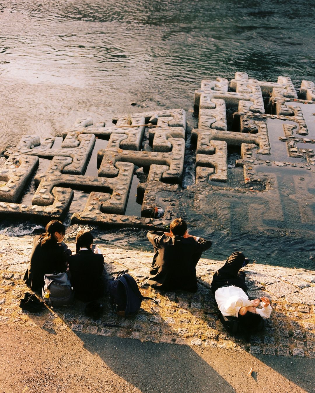 Five people sit by a river with geometric concrete blocks in the background. People have backpacks. The scene is lit by golden hour sunlight.