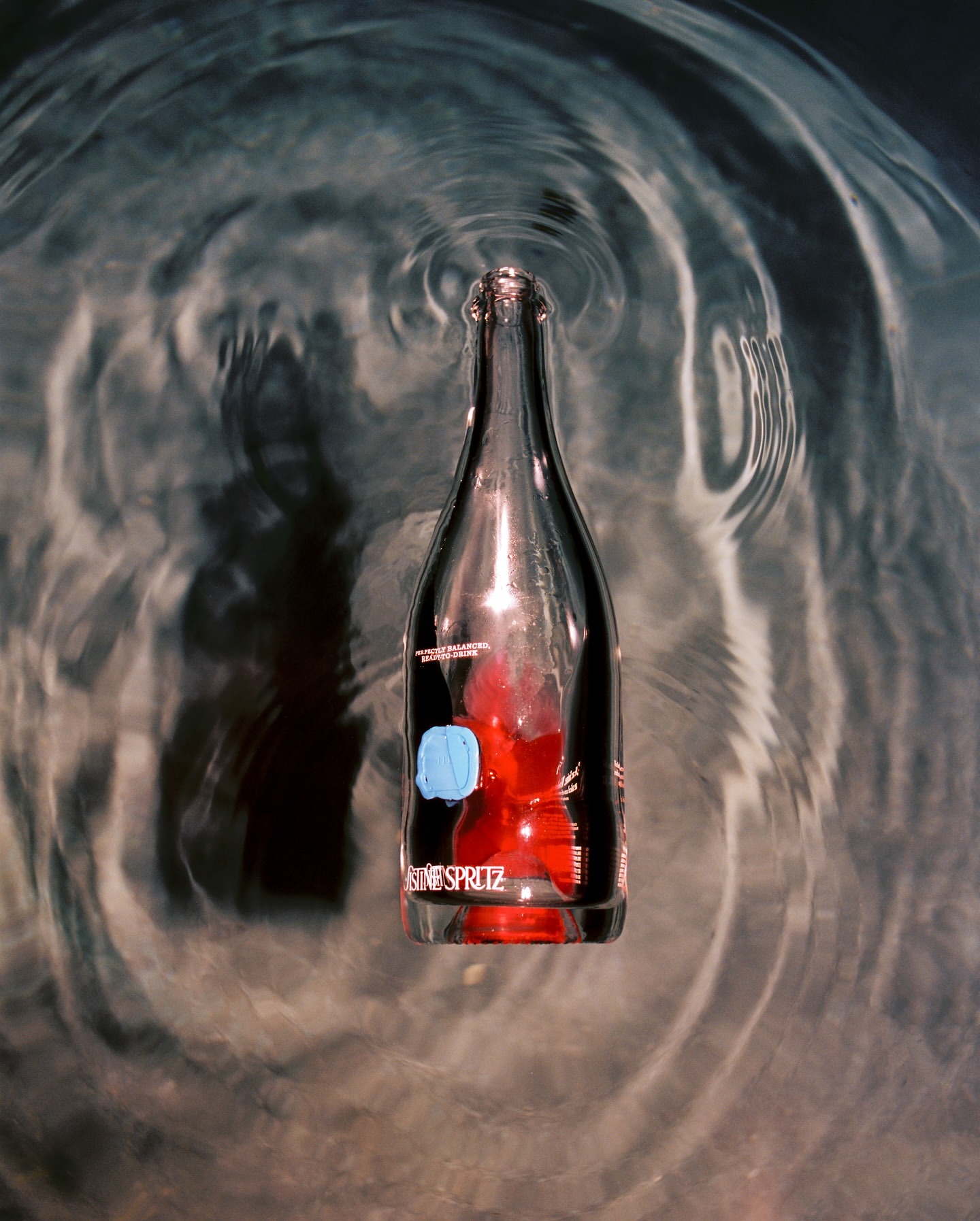 Sparkling glass bottle with red liquid inside floats on water, creating concentric ripples on the surface, top-down perspective.
