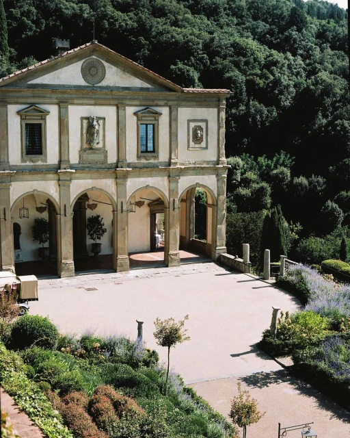 Ornate building with arched openings and stone accents in a lush garden setting with mountains visible in the background on a sunny day.