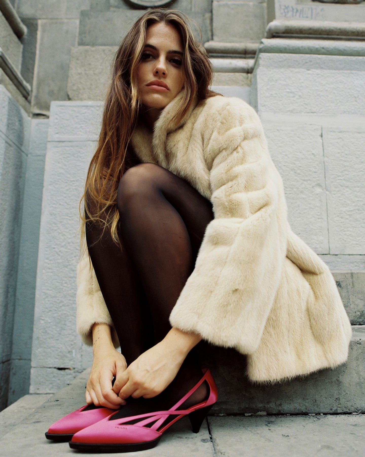 Woman in fur coat sits on grey stone stairs wearing pink heeled shoes and black tights. Long brown hair frames her face in an urban setting.