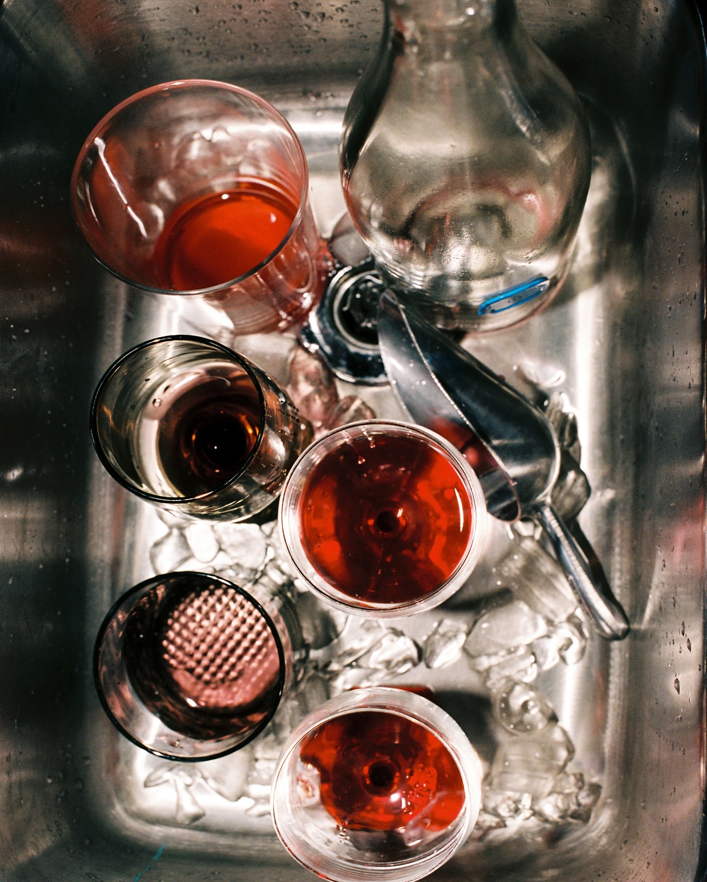 Overhead shot of an ice-filled stainless steel sink holding a glass pitcher, a metal ice scoop, and four glasses containing red and dark liquid.