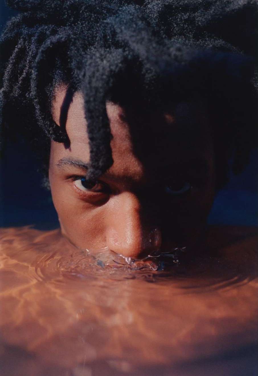 Close-up portrait of a Black man submerged in water with ripples around his nose and mouth, by Pía Riverola.
