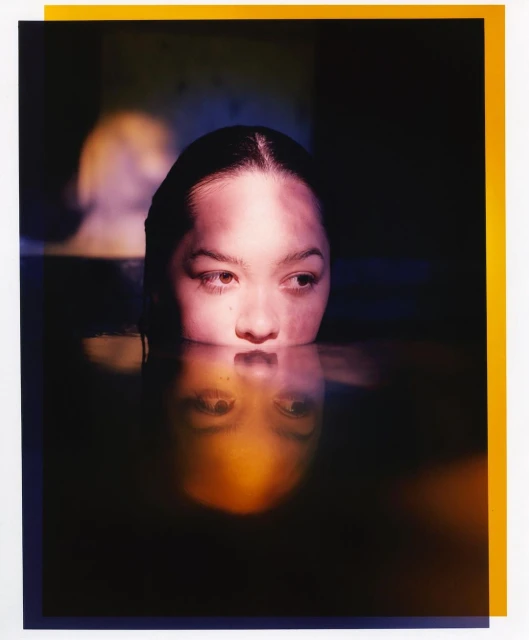 Portrait of a young adult woman with wet hair, partially submerged in water, reflecting with a colorful, dark photo studio background.
