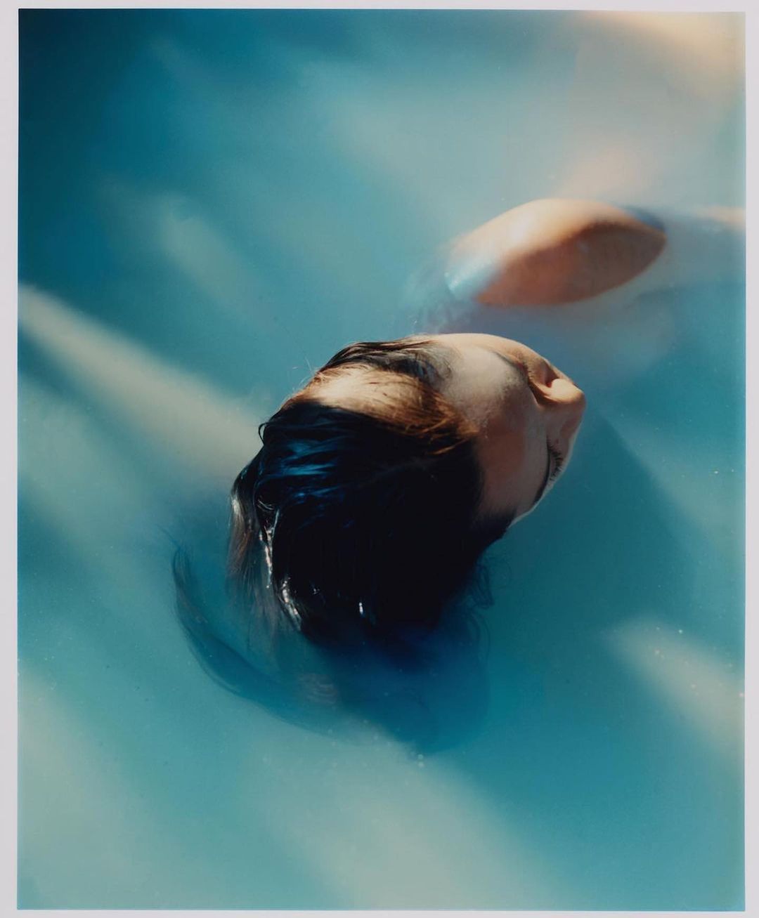 Underwater portrait of an adult female with dark hair partially submerged in light blue water with diffused lighting. Dreamy and surreal.
