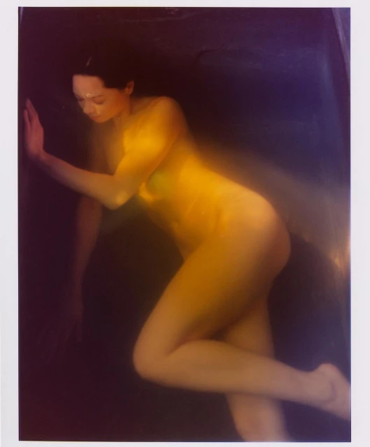 Artistic nude portrait of an adult woman in an amber-toned, dark studio setting, showcasing fine art photography with a blurred effect.
