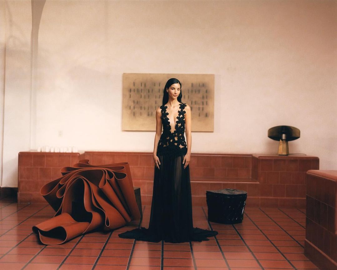Woman in black gown poses in an interior space with terracotta tile floors and decorative art; elegant fashion editorial, modern design.