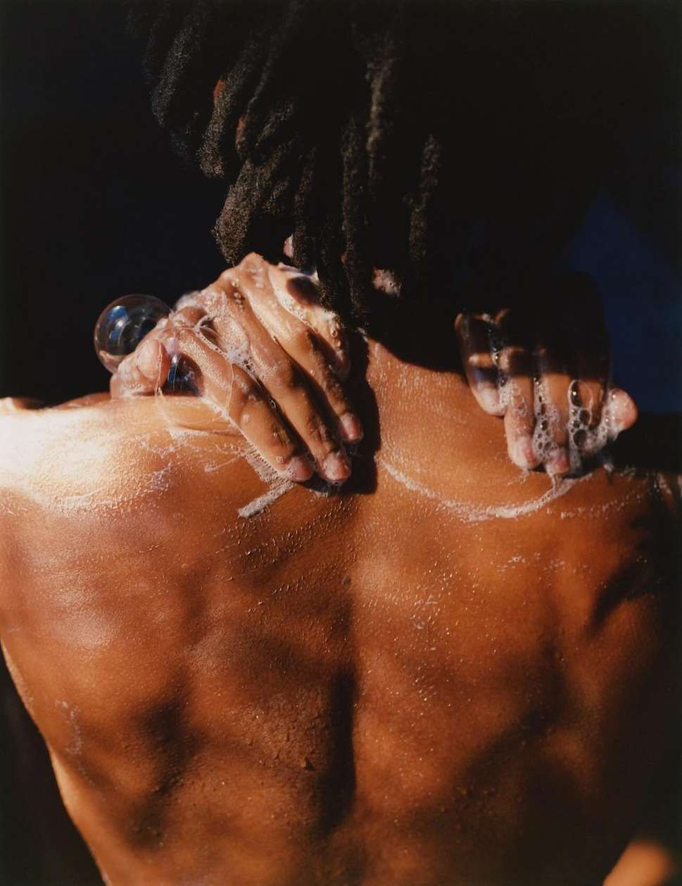 Person washes their back and shoulders with soap, creating suds, dark hair, and brown skin against a dark blue and black background.