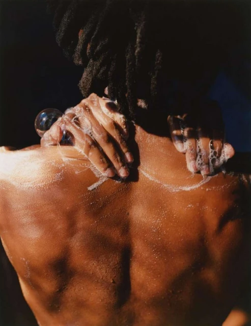 Person washes their back and shoulders with soap, creating suds, dark hair, and brown skin against a dark blue and black background.