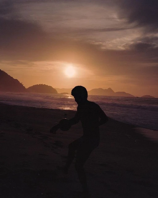 Silhouette of adult male playing with ball on beach at sunset with ocean, islands and cloudscape in background in warm tones.