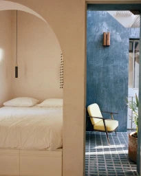 Interior view through an arched doorway of a bedroom with white bedding and a courtyard with a yellow armchair