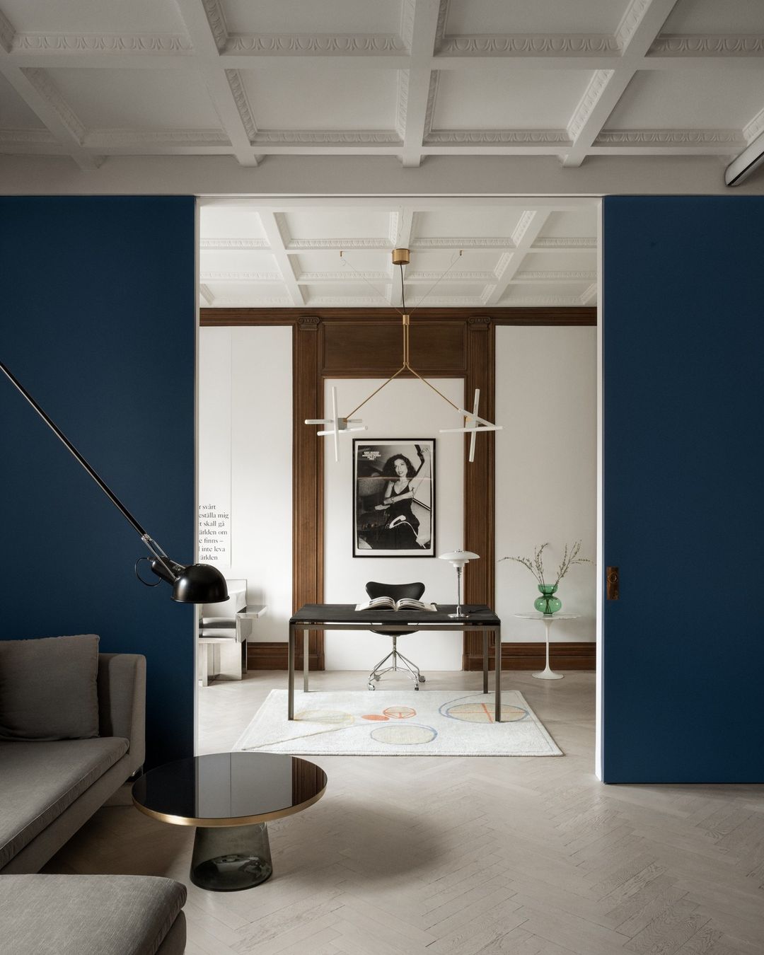 Interior view of a modern home office area featuring a desk, chair, and a contemporary lamp with artwork and gray couch.