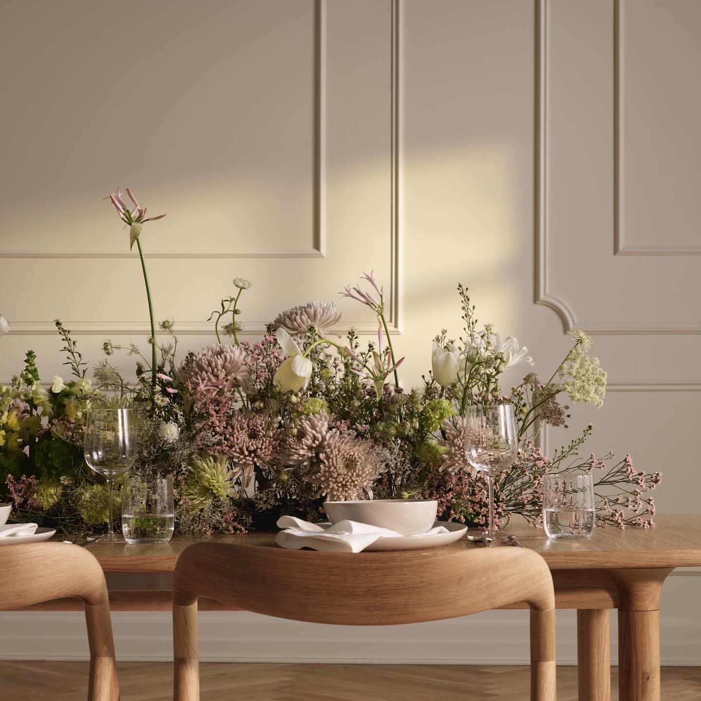 Elegant dining setup featuring a wooden table adorned with a lush floral arrangement, glassware, and plates against a neutral background.