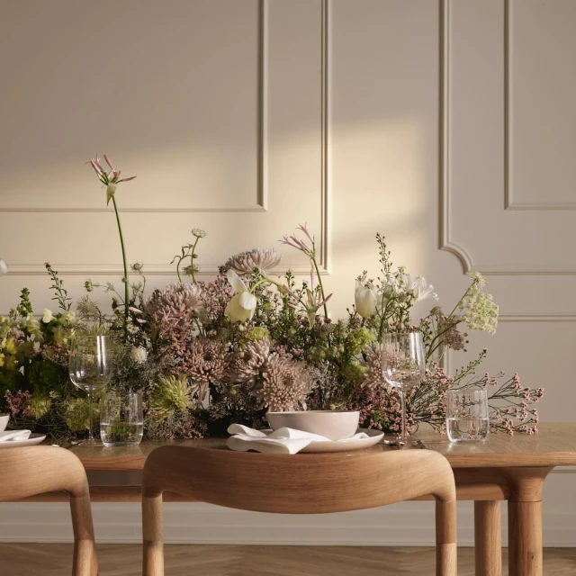 Elegant dining setup featuring a wooden table adorned with a lush floral arrangement, glassware, and plates against a neutral background.