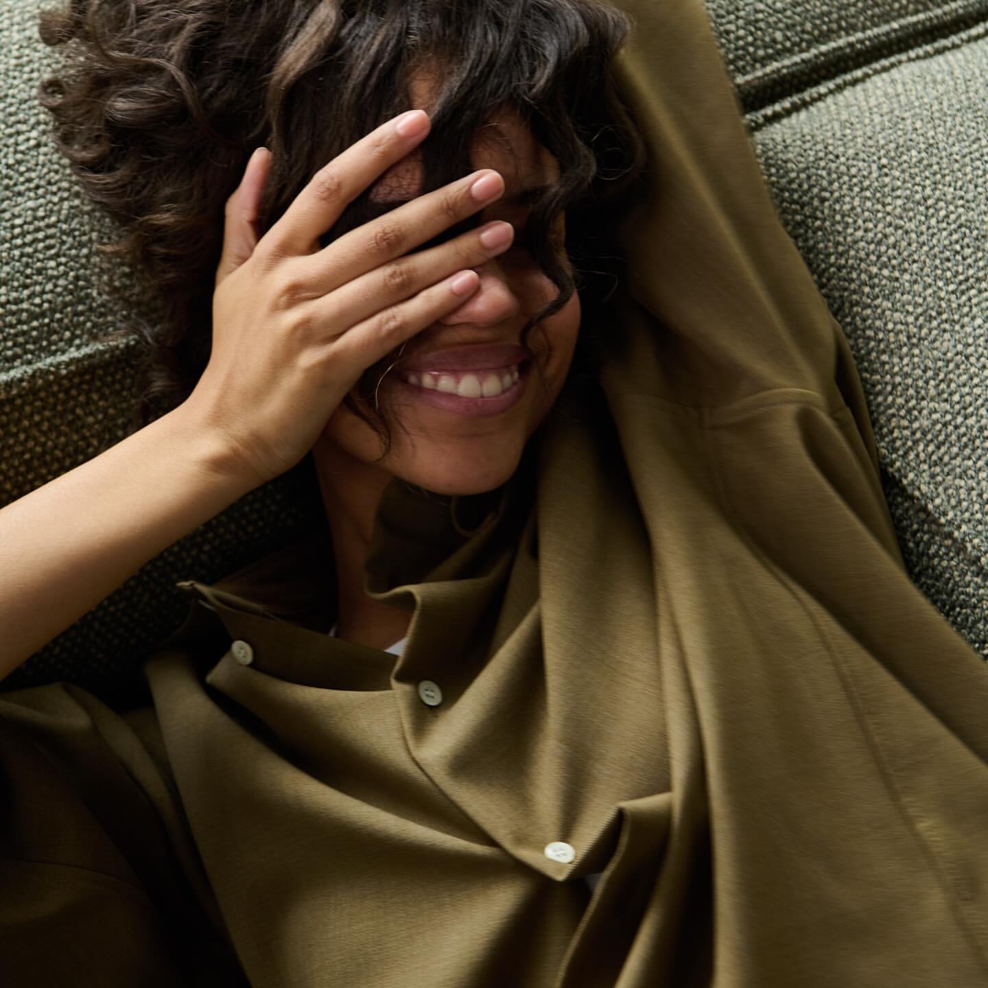 Smiling woman with dark curly hair covers one eye with her hand while relaxing on a green textured sofa wearing an olive-green shirt.