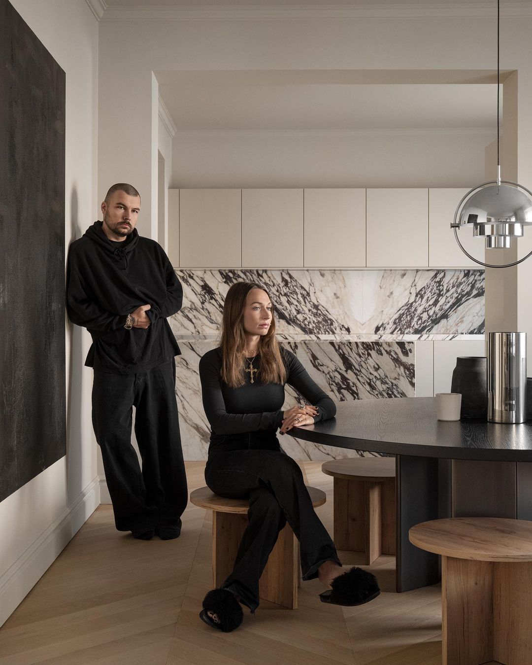 Two adults in an interior setting with wood flooring, a round table, and modern marble backsplash. A painting hangs on the wall.
