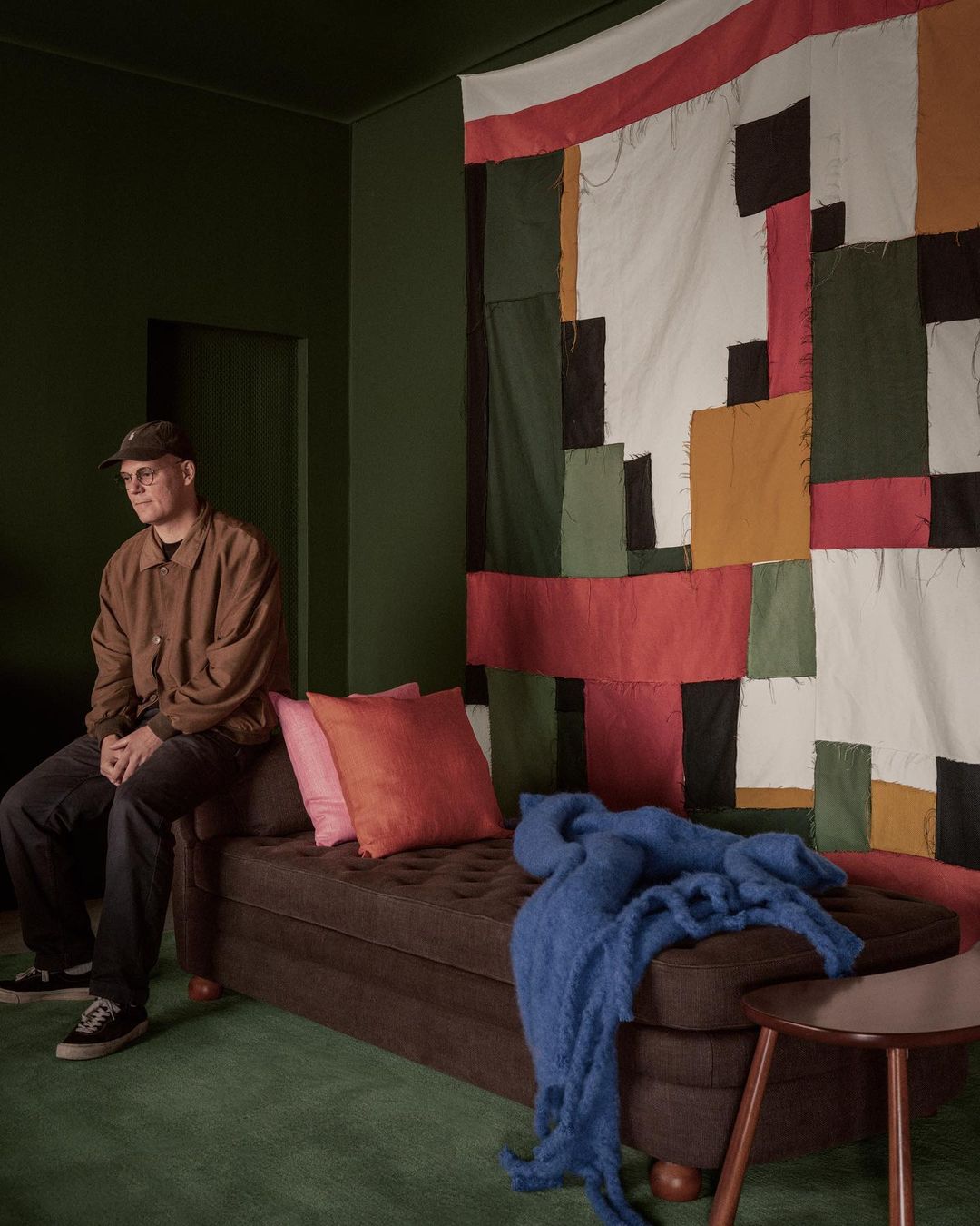 A man sits on a brown daybed in a room with dark green walls and carpet, featuring a colorful patchwork quilt wall hanging.