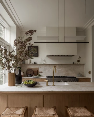 Stylish kitchen interior featuring a marble countertop island with woven stools, a gold faucet, and decorative dried foliage.