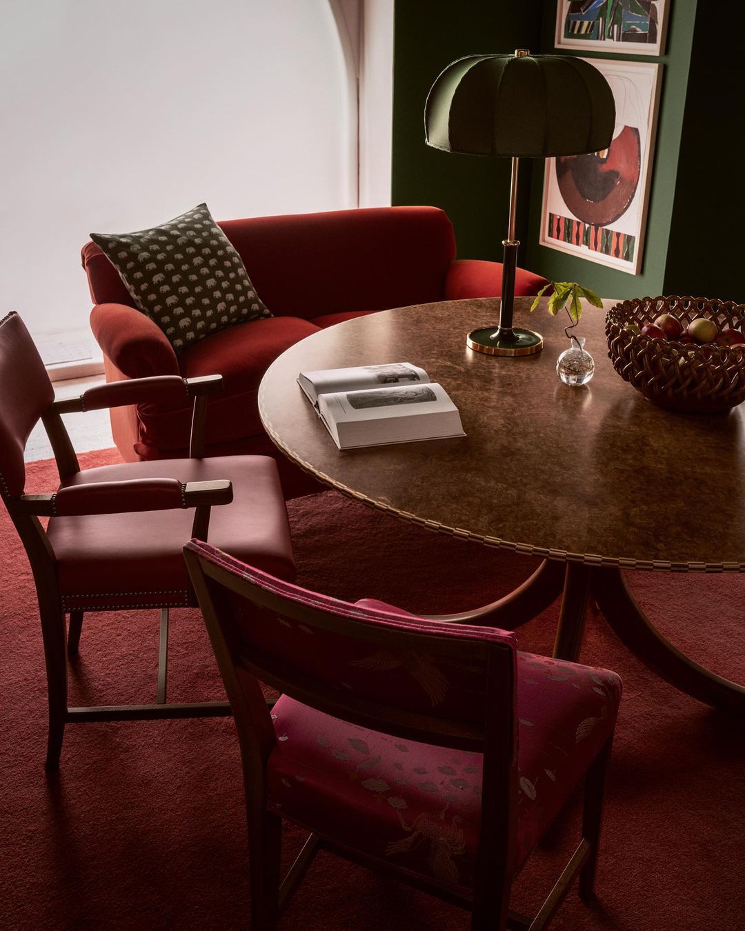 Living room interior featuring a round wood table, two red chairs, a red loveseat, and an olive green lamp against a dark green wall.