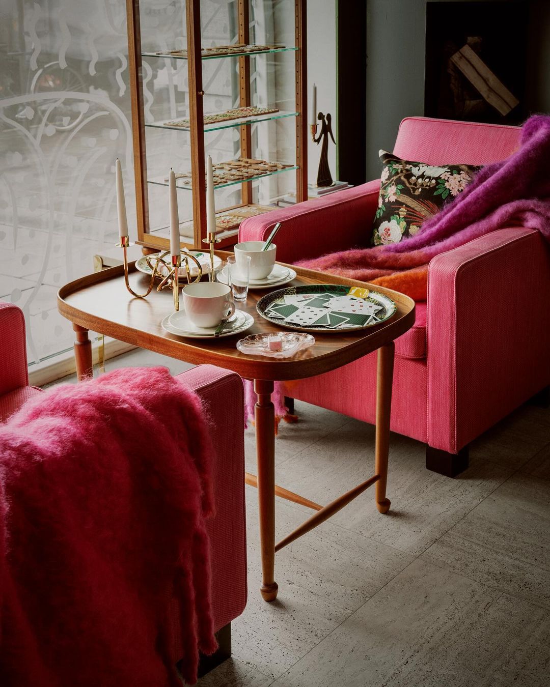 Living room interior featuring pink armchairs, a wooden table with tea set and candles, against a window and display cabinet.