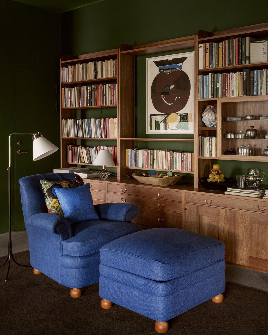 Interior view of a blue armchair and ottoman in a room with a bookcase filled with books and a dark green wall.
