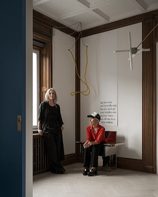 Two adult women indoors in room with white walls, dark wood trim, and minimalist art hanging, with a window with radiator.
