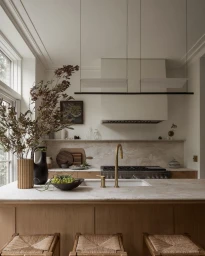 Modern minimalist kitchen interior with marble countertops, wooden island, brass fixtures, and a large dried botanical arrangement.
