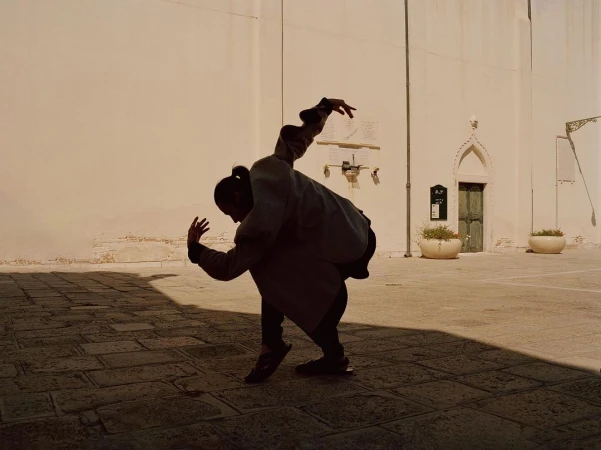 A single person in motion with raised hands and a bent posture captured in an outdoor location in front of a building with a stone wall.