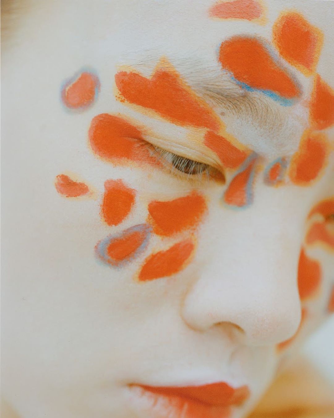 Abstract close-up of a person's face with orange paint on their face and lips in a beauty or fashion-related setting.