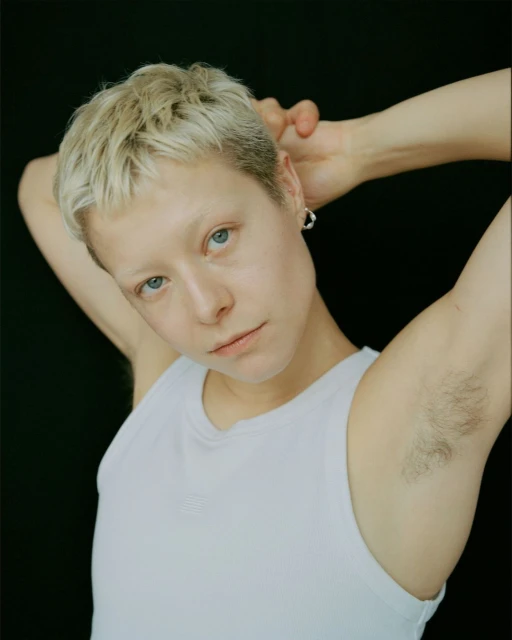 Close-up portrait of a young adult female with a short blonde buzzcut, wearing a white tank top, posing against a dark background.