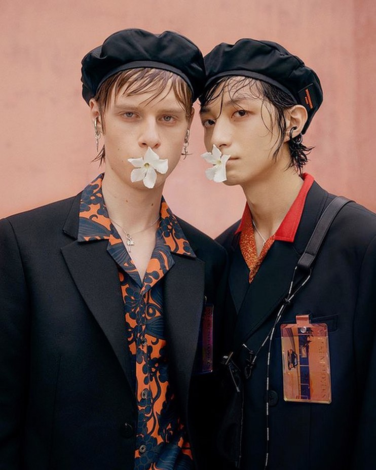 Close-up of two adult male models wearing black blazers, black berets, and holding white flowers in their mouths against a pink backdrop.