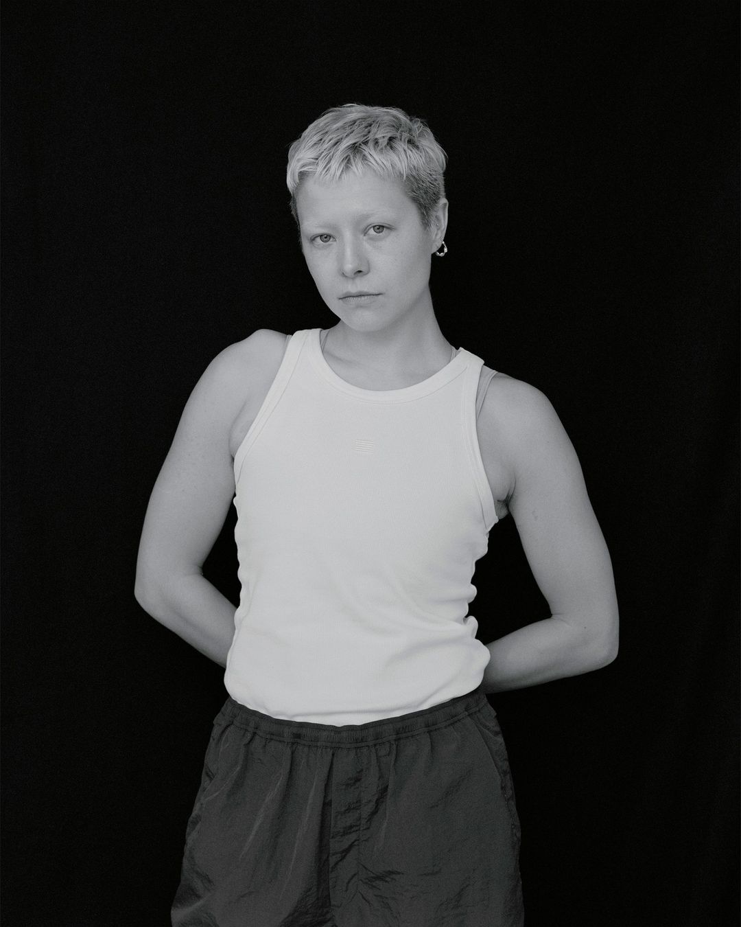 Monochrome portrait of a person with short blonde hair wearing a white tank top and dark shorts, standing against a black backdrop.