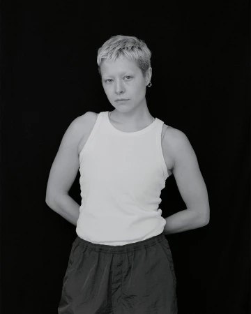 Monochrome portrait of a person with short blonde hair wearing a white tank top and dark shorts, standing against a black backdrop.