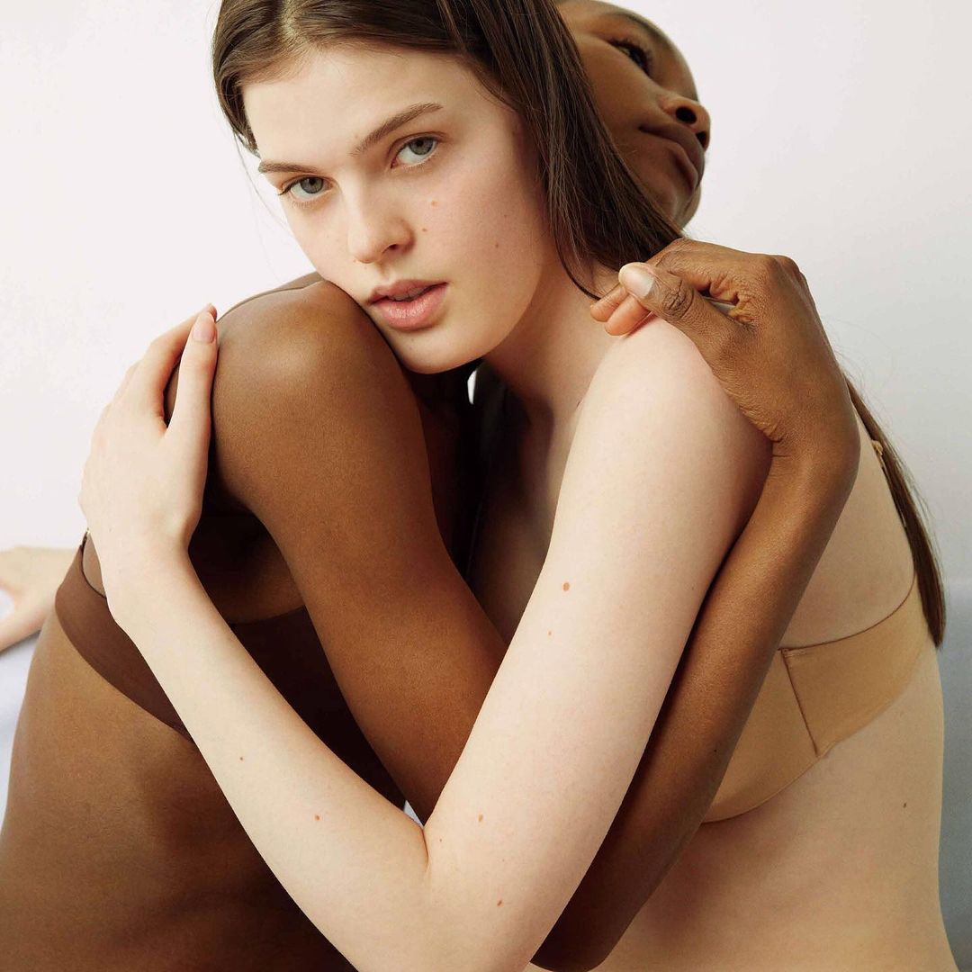 Two women embracing in front of a white background. One woman has fair skin and brown hair, and the other has dark skin.