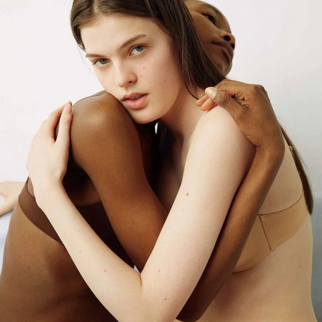 Two women embracing in front of a white background. One woman has fair skin and brown hair, and the other has dark skin.