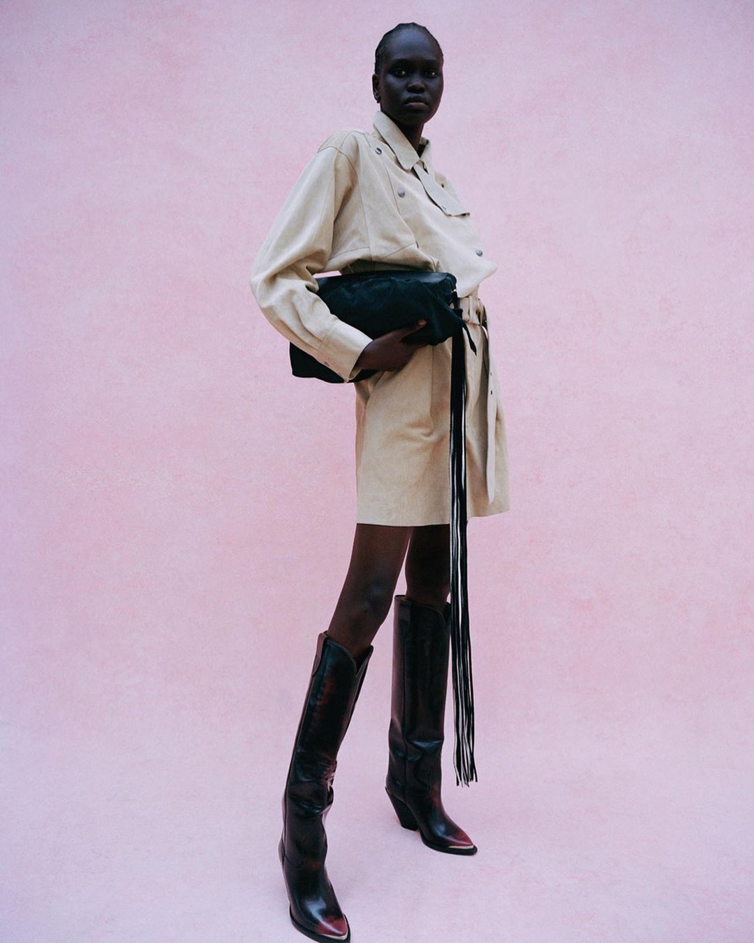 Full-length portrait of an adult female fashion model posing in a studio with a clutch purse, wearing a belted dress and black western boots.